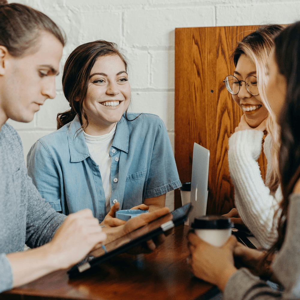 A group of four people appearing to work collaboratively together in a coffee shop setting. One is working on a laptop, one is working on a tablet, and the last two are having a conversation with each other.